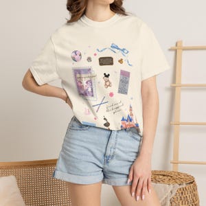 May include: Cream-colored t-shirt featuring a collage of whimsical illustrations. The design includes a purple postage stamp, a teddy bear, a castle, and the text "find me at the happiest place". The shirt is paired with denim shorts.