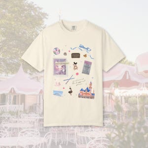 May include: Cream-colored t-shirt featuring a whimsical design with a castle, stamps, and a bow. The shirt has the text "find me at the happiest place". The design includes a Mickey Mouse illustration and other decorative elements.