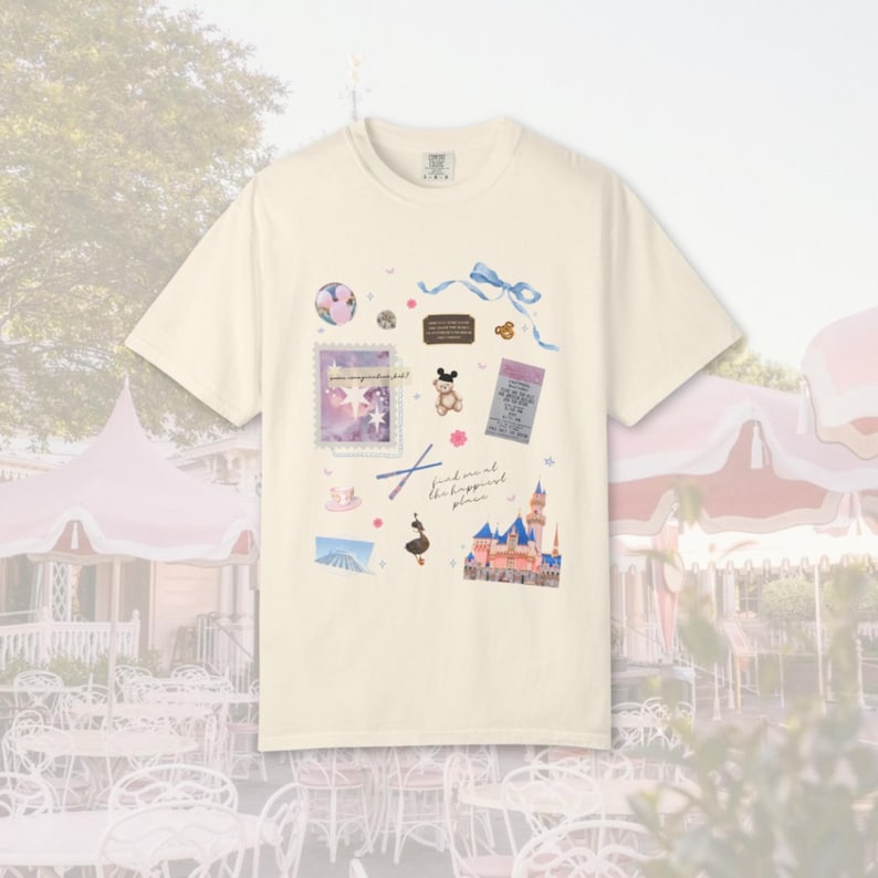 May include: Cream-colored t-shirt featuring a whimsical design with a castle, stamps, and a bow. The shirt has the text "find me at the happiest place". The design includes a Mickey Mouse illustration and other decorative elements.