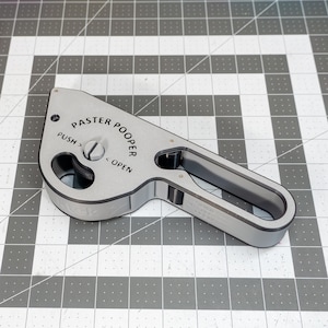 May include: A silver-colored tool with the words "PASTER POOPER" and "PUSH" and "OPEN" printed on it. The tool has a handle and a curved design. The tool is on a gray and white checkered surface.