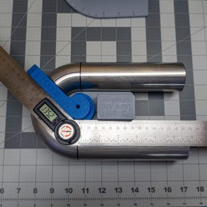 May include: A blue and black digital angle finder measuring a stainless steel pipe with a 13.52 degree angle. The pipe is connected to a straight piece of pipe. The pipe is labeled "WRKTE OD 175 GER 2".