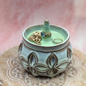 Könnte beinhalten: Ein Keramik-Schmuckhalter mit hellblauem und braunem Blumendesign. Der Innenraum ist hellgrün und enthält Ringe und eine goldene Halskette. Der Ringhalter hat eine konische Form. Der Halter steht auf einer Holzoberfläche.