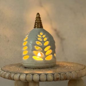 Puede incluir: Un portavelas de cerámica gris con recortes en forma de hojas, rematado con un adorno dorado. El portavelas se encuentra sobre una mesa de madera decorativa. La vela en el interior emite un brillo cálido y amarillo.