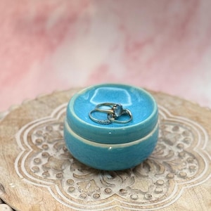 May include: A small, round, turquoise ceramic jewellery box with a lid, holding three rings. The box sits on a decorative wooden tray with a carved floral pattern. The background is a soft pink.