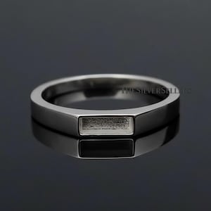 Op de afbeelding: Een zilveren ring met een rechthoekige zetting voor een steen. De ring heeft een gepolijste afwerking en een minimalistisch ontwerp. De ring wordt gepresenteerd op een donker, reflecterend oppervlak.