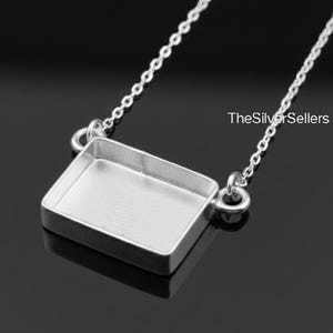 Pode incluir: Um colar prateado com um pingente quadrado. O pingente é conectado a uma corrente fina. A imagem é definida contra um fundo escuro, destacando as qualidades reflexivas do metal. O texto "TheSilverSellers" é visível.