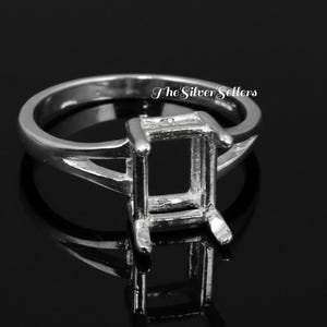 Puede incluir: Un anillo de plata con un engaste cuadrado, diseñado para sostener una piedra preciosa. El anillo tiene una caña dividida y un acabado pulido. El texto "The Silver Sellers" es visible en el anillo.