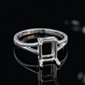 Pode incluir: Um engaste de anel em metal prateado polido com uma cabeça retangular de quatro garras projetada para segurar uma pedra preciosa. A banda tem um design de haste dividida que se estreita em direção ao centro, criando uma aparência limpa e estruturada.