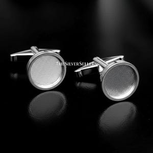 May include: Two silver cufflinks with a circular design and a textured, striped pattern. The cufflinks have a shiny finish and a T-shaped post for securing them to a shirt cuff. The background is black.