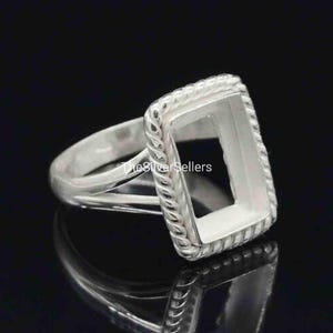 Op de afbeelding: Een zilveren ring met een rechthoekige zetting. De ring heeft een gedraaid touwontwerp rond de rand van de zetting. De ring wordt getoond op een reflecterend zwart oppervlak.