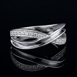 May include: A silver-toned ring featuring a multi-layered crossover design. One band is encrusted with a row of small, clear, sparkling stones, while the other bands remain smooth and polished, creating an intertwined, fluid appearance.
