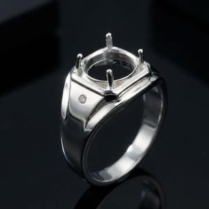 May include: A polished silver-tone ring setting featuring a square-shaped head with four prongs designed to hold a center stone. The band includes a single small, round, clear accent stone embedded directly into the metal shank below the head.