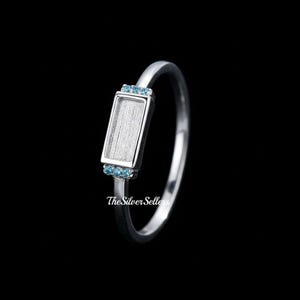 Op de afbeelding: Een zilveren ring met een rechthoekige zetting, versierd met kleine blauwe edelstenen. De ring heeft een strak, modern design, perfect voor het presenteren van een op maat gemaakte steen of ontwerp. Op zwarte achtergrond.