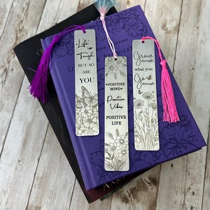 May include: Three silver bookmark with floral designs and inspirational quotes. The bookmarks have pink, purple, and blue tassels. The quotes are "Life is tough but so are you", "Positive Mind Positive Vibes", and "Grow through what you go through".