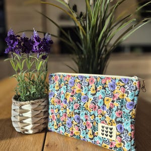 Op de afbeelding: Een rechthoekig gewatteerd tasje met een levendig bloemenpatroon in blauw, geel, roze en zwart. Het tasje heeft een crèmekleurige rits en een klein rechthoekig label aan de voorzijde met een patroon van negen zwarte hartjes.