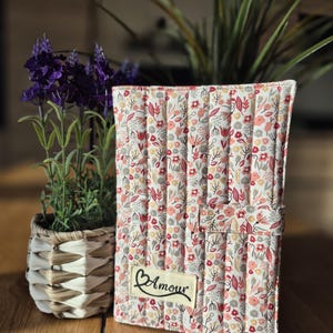 Può includere: Copertina per quaderno in tessuto trapuntato con motivo floreale rosa, rosso e grigio su sfondo bianco. La copertina ha un'etichetta con la scritta "Amour" in corsivo. Il quaderno è accanto a una piccola pianta in vaso con fiori viola.