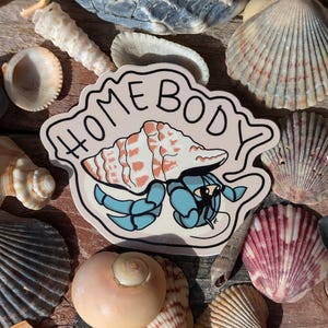 May include: A sticker with a pink background and the text "Homebody" in black. A blue hermit crab is drawn on the sticker, carrying a pink and white seashell on its back.