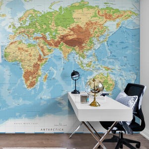 May include: A large world map mural in shades of blue, green, brown, and tan covers a wall. A white desk with a black chair and decorative globes are in front of the map. A floor lamp with a white shade is on the left.