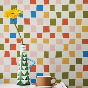 May include: A still life featuring a yellow gerbera daisy in a green and white vase with a blue handle, a brown coffee cup and saucer, and a white nightstand. The background is a repeating pattern of colourful squares.