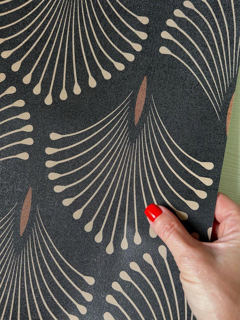 May include: Close-up of a black fabric with a repeating pattern of beige fan-like shapes and brown elongated ovals. The fabric is being held by a hand with red nail polish.