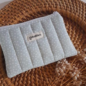 以下が含まれることがあります： 白のドット柄が入った、水色のキルティングポーチ。ファスナー開閉で、「Mollies」のラベル付き。編まれた柳のバスケットの上に置かれ、ドライフラワーが添えられています。小物の収納に最適です。