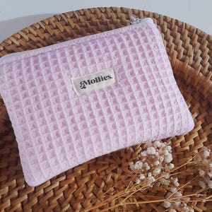 Peut inclure: Une pochette rose clair à texture gaufrée avec une fermeture éclair. La pochette porte une étiquette blanche rectangulaire avec le texte "Mollies". Elle repose sur un panier tressé avec des fleurs séchées.