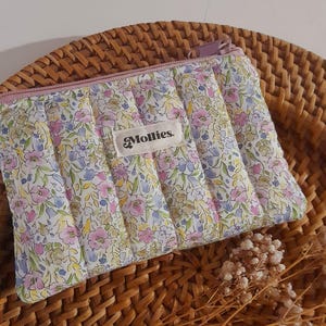 以下が含まれることがあります： ピンク色のファスナーと「Mollies」のブランド名が小さなラベルに付いた、キルティング加工された花柄のポーチ。 ピンク、青、黄色の小花模様が特徴です。 ポーチは、編まれた籐の表面に置かれています。