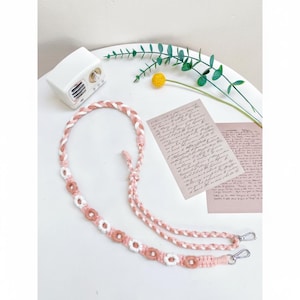 Op de afbeelding: Een roze en witte gevlochten lanyard met een bloemenontwerp. De lanyard heeft zilverkleurige metalen sluitingen en is versierd met kleine, gehaakte bloemen. De achtergrond bevat een witte radio, handgeschreven notities en decoratief groen.