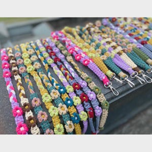 May include: A collection of colorful macrame lanyards with floral and braided designs. The lanyards feature various colors, including pink, yellow, green, and purple, and are adorned with small flower accents and metal clasps.