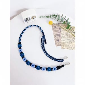 Op de afbeelding: Een blauw-witte gevlochten riem met zilveren metalen sluitingen. De riem is versierd met kleine, blauwe gehaakte bloemen. De riem is ontworpen voor een tas of portemonnee en wordt gepresenteerd op een wit oppervlak met decoratieve elementen.