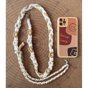 May include: A macrame phone strap in cream and tan with flower accents and a silver clasp. A phone case with a patchwork design, a smiley face, and the words "Good Luck" is next to the strap.