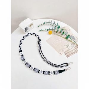 Op de afbeelding: Een handgemaakte, gevlochten telefoonriem in zwart, wit en grijs. De riem heeft een bloemenontwerp met kleine witte en grijze bloemen. Het heeft zilverkleurige metalen sluitingen voor eenvoudige bevestiging aan een telefoonhoesje.