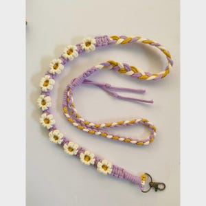 Op de afbeelding: Een handgemaakte macramé lanyard in tinten lavendel, geel en crème. De lanyard heeft een madeliefjeskettingontwerp met crèmekleurige bloemen en een gevlochten gedeelte. Het heeft een metalen sluiting.