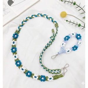 May include: A handmade crochet strap and wristlet set. The strap is green, white, and blue, with a braided design and flower accents. The wristlet is light blue and white, with flower accents and a silver clasp. The set is on a white background.
