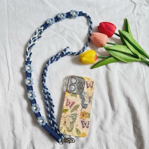 May include: A blue and white braided phone lanyard with light blue flower accents. A phone case with a butterfly pattern is attached. Three tulips in red, pink, and yellow are in the background.