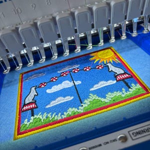 May include: An embroidery machine in action, creating a vibrant patch. The patch depicts a blue sky with clouds, a sun, and two white figures on pedestals. The design is framed with red and yellow borders.