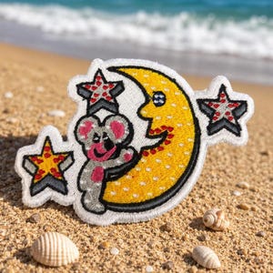 May include: An embroidered patch featuring a grey koala hugging a yellow crescent moon with a face. The design includes three stars with red and white patterns, all outlined in white thread against a textured, white-bordered background.