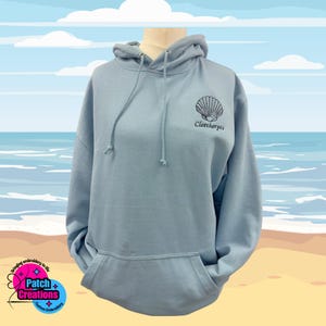 May include: Pale blue hoodie with a drawstring hood and a front pocket. It has an embroidered seashell design and the word "Cleethorpes" in black. The background depicts a beach scene with a blue sky and clouds.