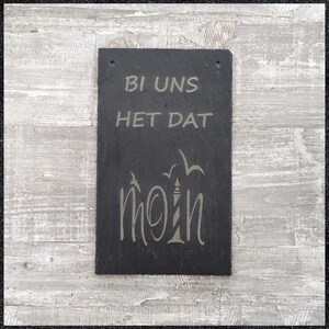 Könnte beinhalten: Schwarzes Schiefer-Schild mit dem Text "Bi uns het dat" und dem Wort "moin" in einer stilisierten Schrift mit einem Leuchtturm und Palmen.