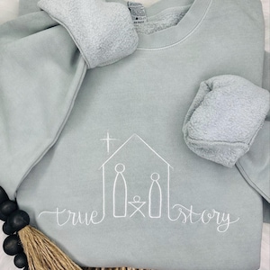 May include: A light blue sweatshirt with a white embroidered design of a manger scene with the words "true story".