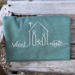 May include: A green sweatshirt with the words "silent night" in cursive. A white line drawing of a nativity scene is above the text. The image is on a wooden surface.