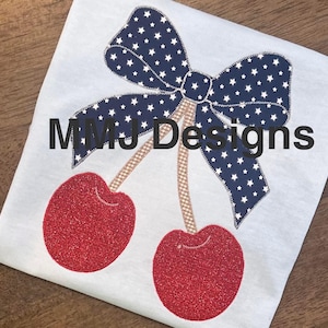 May include: White t-shirt featuring a navy blue bow with white stars and two red glitter cherries. The text "MMJ Designs" is printed in black across the shirt.