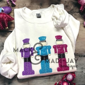 May include: White sweatshirt with three embroidered nutcracker figures. The nutcrackers are wearing purple, blue, and pink sequined outfits. The text "Mad-Jay Boutique" is visible in the bottom right corner.