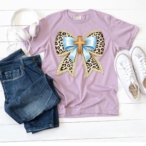 Könnte beinhalten: Ein lilafarbenes T-Shirt mit einem großen Schleifendesign mit Leopardenmuster, einem hellblauen Band und einem goldenen Kreuz in der Mitte. Das Bild zeigt auch blaue Jeans, weiße Turnschuhe und weiße Kopfhörer.