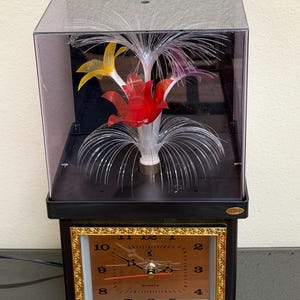 Puede incluir: Un reloj de cuarzo negro con marco dorado y esfera cuadrada con números y manecillas negras. Sobre el reloj hay una vitrina de acrílico transparente que contiene una lámpara de fibra óptica con detalles florales de plástico rojo, amarillo y morado.