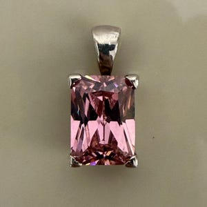 May include: A rectangular, radiant-cut pink gemstone pendant set in a silver-toned metal prong setting. The pendant features a polished, reflective silver bail at the top, designed for attachment to a chain, showcasing a clear, faceted pink stone.