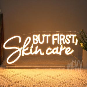 May include: A neon sign with the words "BUT FIRST, Skin care" in a cursive font. The sign is lit with a warm, white glow and is mounted on a clear backing. The sign is placed on a table with a patterned tablecloth.