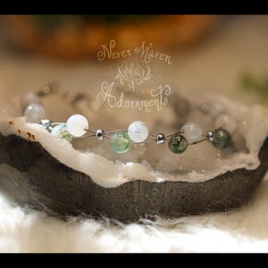 May include: A delicate bracelet featuring white and green gemstone beads strung on a silver wire. The bracelet rests inside a geode, with the text "Never Maren Adornments" in the background.