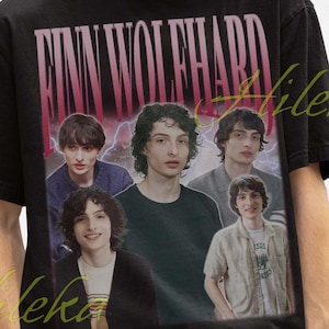 May include: Black t-shirt featuring a graphic of Finn Wolfhard and the text "Finn Wolfhard" in a pink font. The shirt has a vintage aesthetic. The person in the image is wearing a black bucket hat.