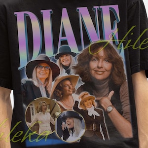 Puede incluir: Camiseta negra con un gráfico de Diane Keaton, con el nombre "DIANE" en una fuente degradada. El diseño incluye múltiples imágenes de la actriz. La camiseta la lleva una persona con un sombrero de pescador negro.